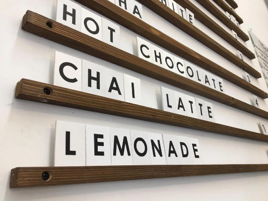 Ahşap çıtalı duvar menü panosu, beyaz PVC harflerle yazılmış fiyat listesi.