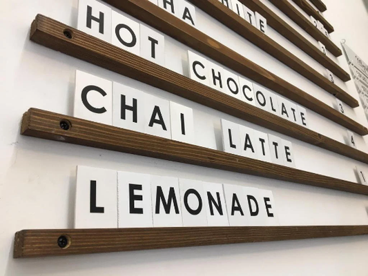 Ahşap çıtalı duvar menü panosu, beyaz PVC harflerle yazılmış fiyat listesi.