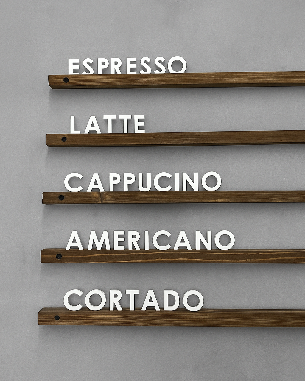 Gri duvar zemininde, koyu ahşap raflara yerleştirilmiş beyaz harflerle yazılmış minimalist kahve menüsü: Espresso, Latte, Cappuccino, Americano ve Cortado.